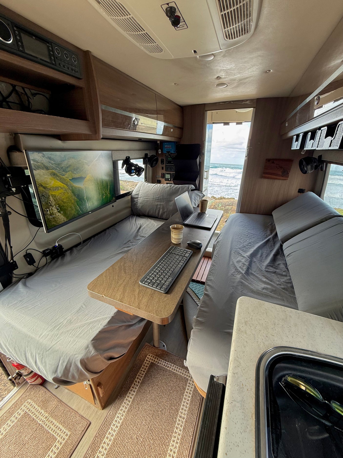 Cozy RV rental workspace with beds, a monitor, laptop, and scenic ocean views from the open rear.