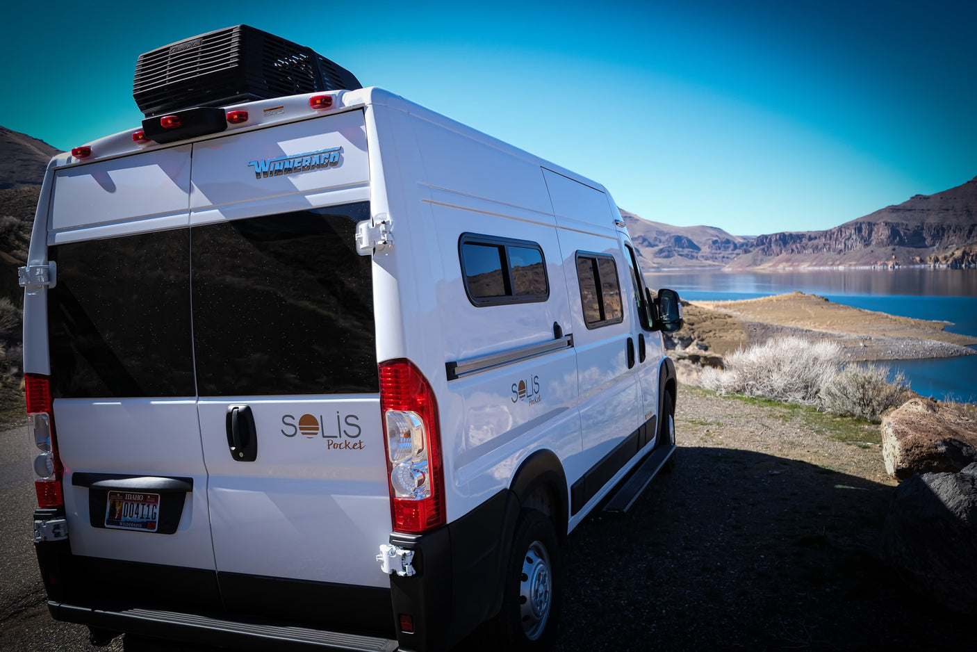 White Winnebago Solis Pocket camper van, ideal for rv rental, parked by a scenic lake with mountains under a blue sky.
