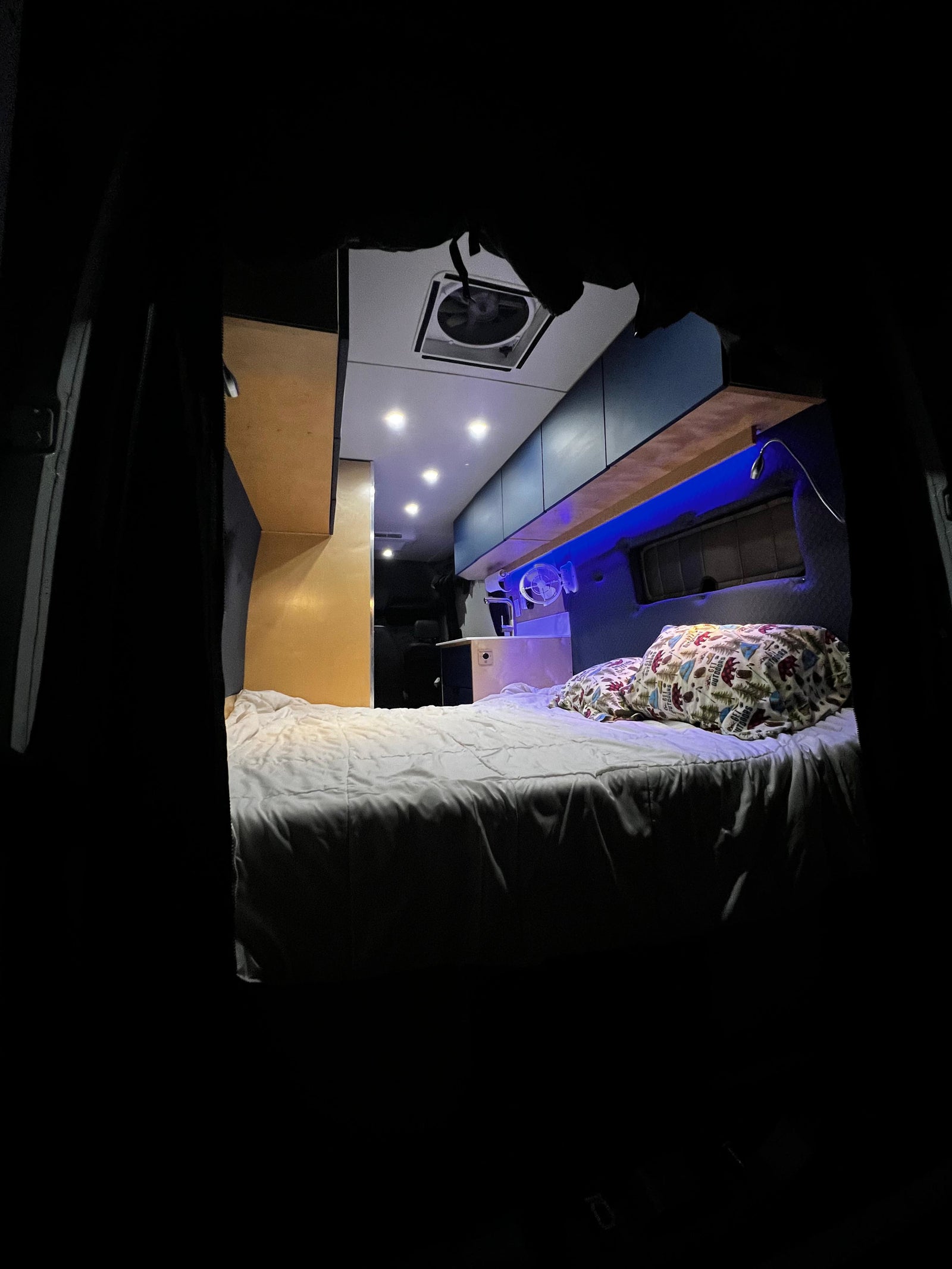 Inside this rv rental, a cozy bed with patterned pillows, blue accent lighting, overhead spotlights, and a ceiling fan create comfort.