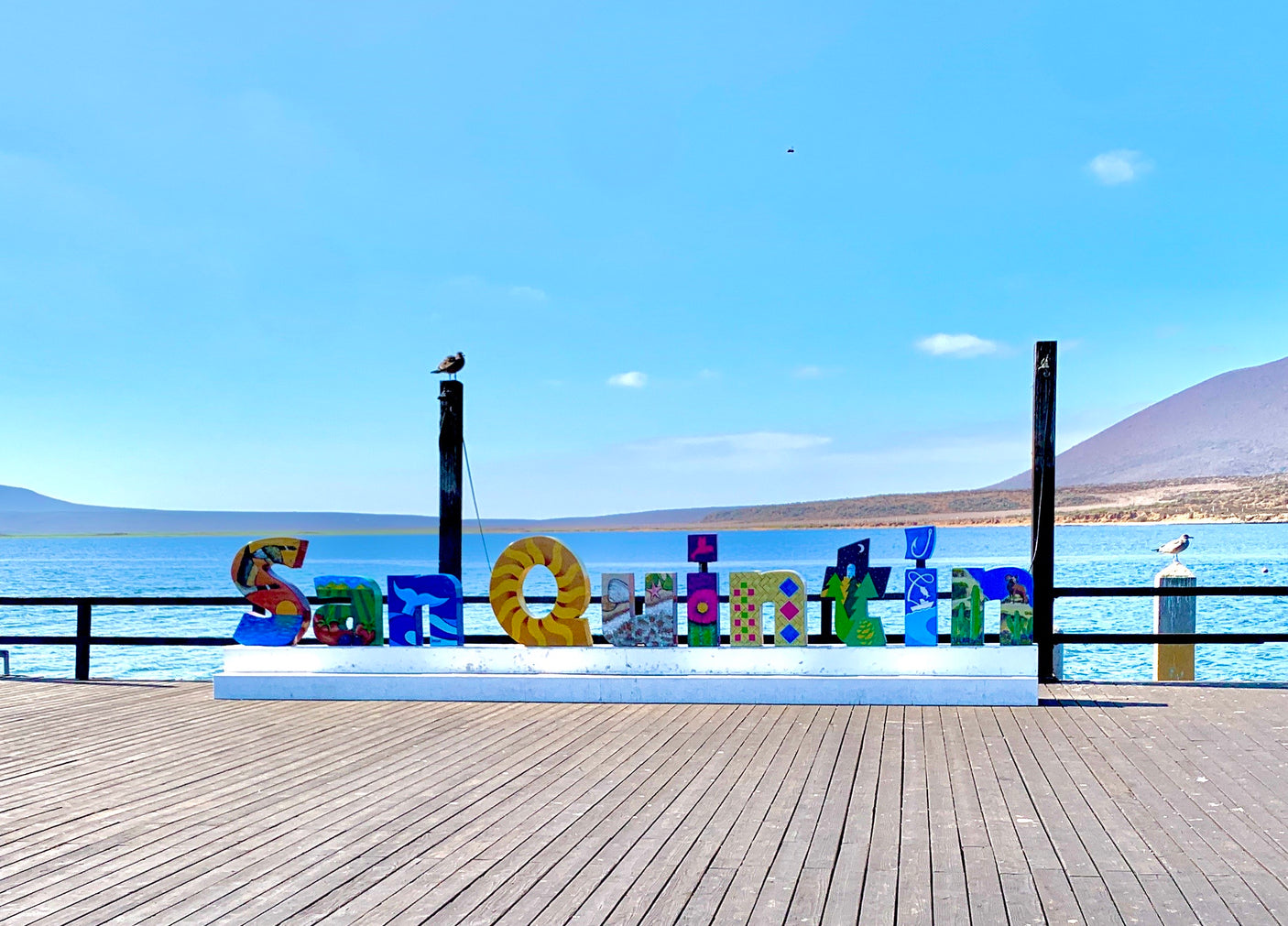 Colorful "San Quintin" sign on a waterfront pier, under a clear blue sky with distant mountains. Perfect for an RV rental getaway, with gulls resting nearby.