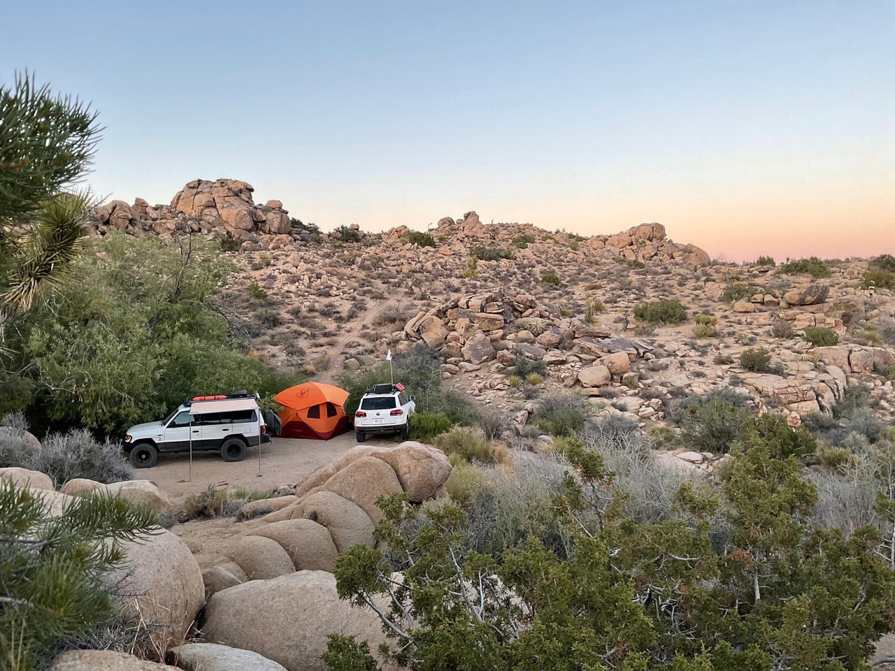 Desert camping setup: white SUVs, orange tent among large rocks. Plan your rugged RV rental adventure in this landscape.