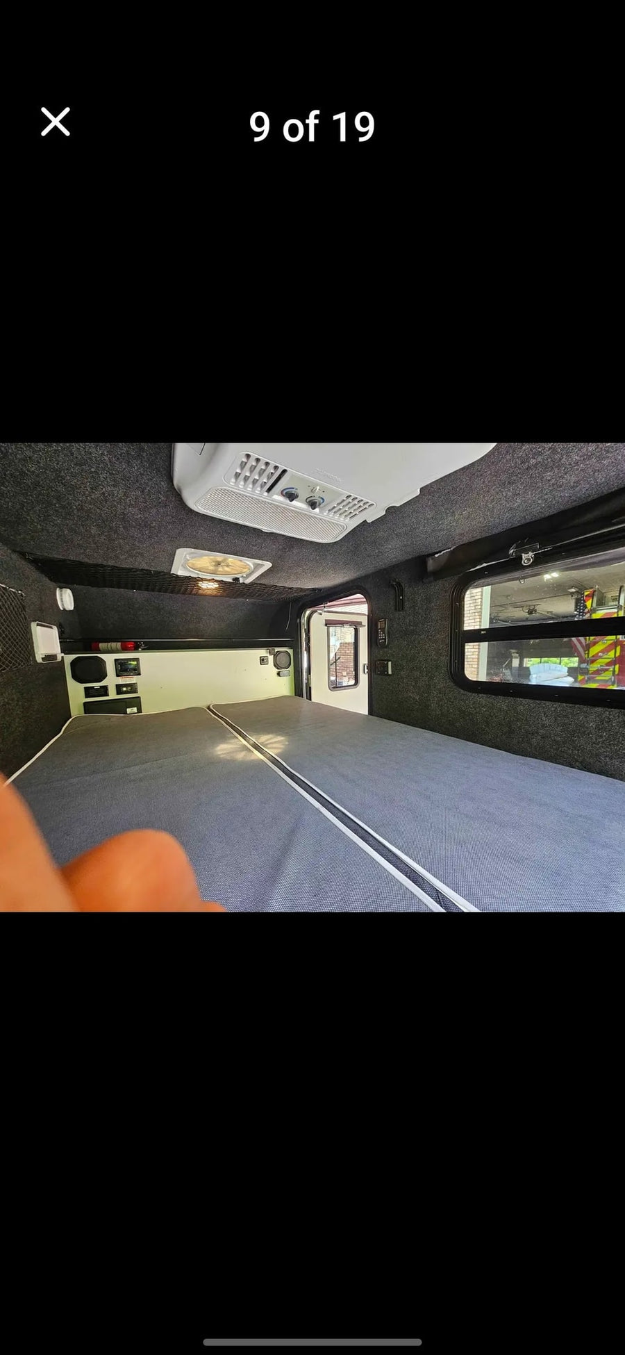 Inside this RV rental: a spacious gray bed, dark upholstered interior with AC, fan, controls, and a window view.