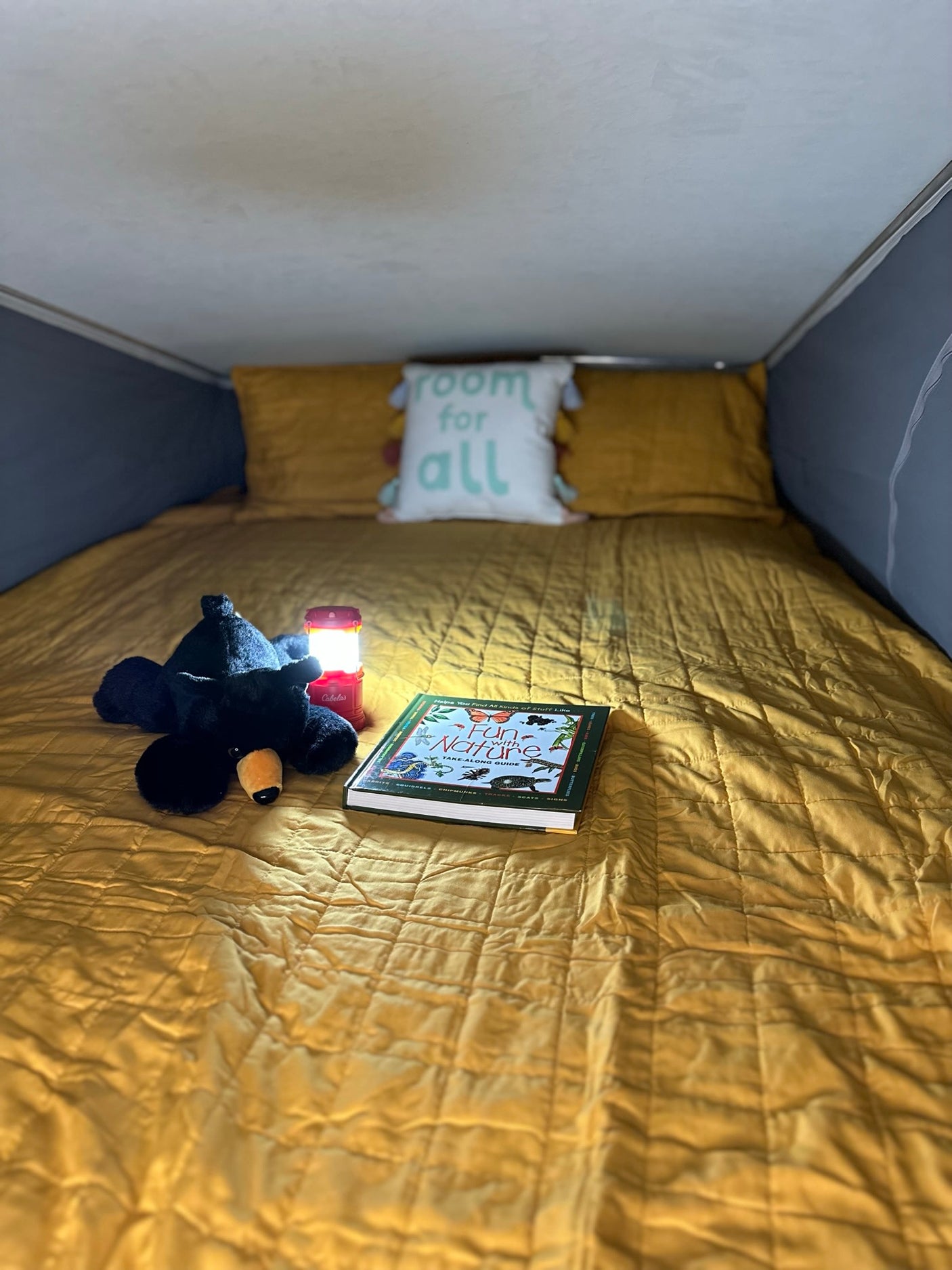Cozy RV rental bed with yellow quilt, plush bear, lit lantern, and a 'Fun with Nature' book. Perfect for reading on camping adventures.