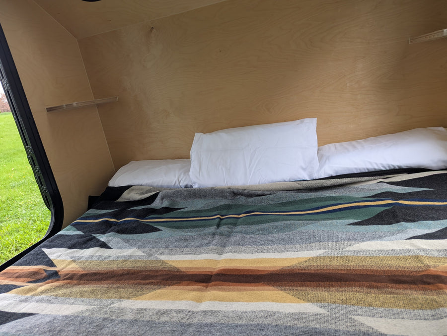 Cozy RV rental interior shows a sleeping area with wooden walls, a small shelf, pillows, and a vibrant patterned blanket.