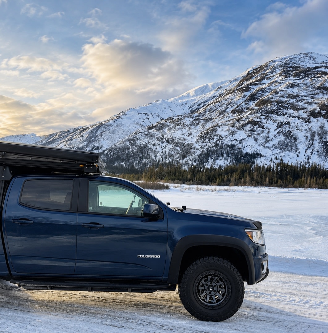 This blue truck camper setup, ideal for an RV rental, is parked in a snowy mountain landscape by a frozen lake.