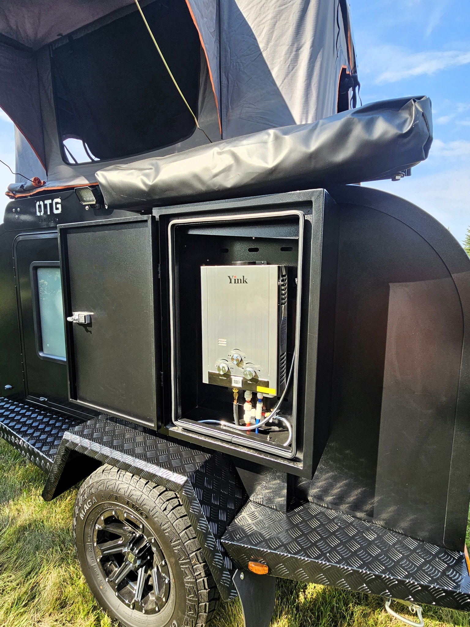 ready for adventure.|Black OTG teardrop RV rental featuring an external Yink water heater and rooftop tent. Ready for adventure.|View inside an RV rental's rear hatch revealing a black outdoor kitchen: pull-out stove