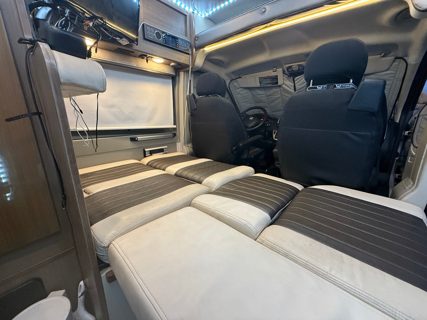 RV rental: Inside a camper van, seats convert to a spacious bed with white & grey cushions. Driver's area is visible.