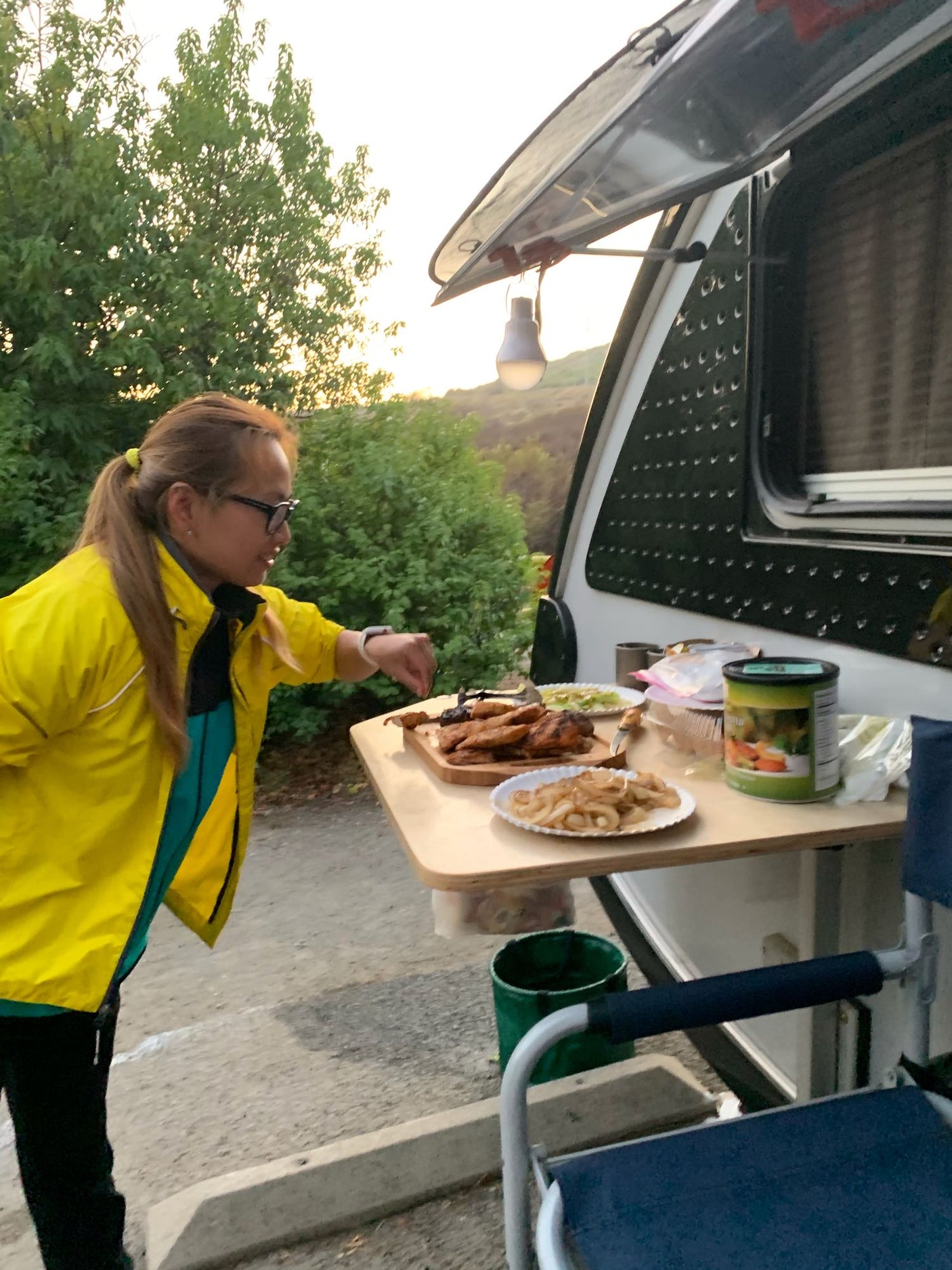 Woman prepares dinner at her RV rental campsite, featuring grilled chicken and onions on a table.