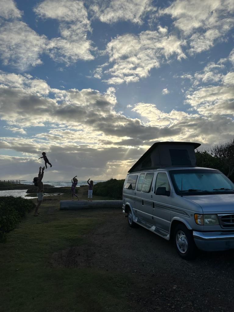 Family joy at sunset, tossing kids by the sea next to a parked RV rental. A silver pop-top van and cloudy sky.
