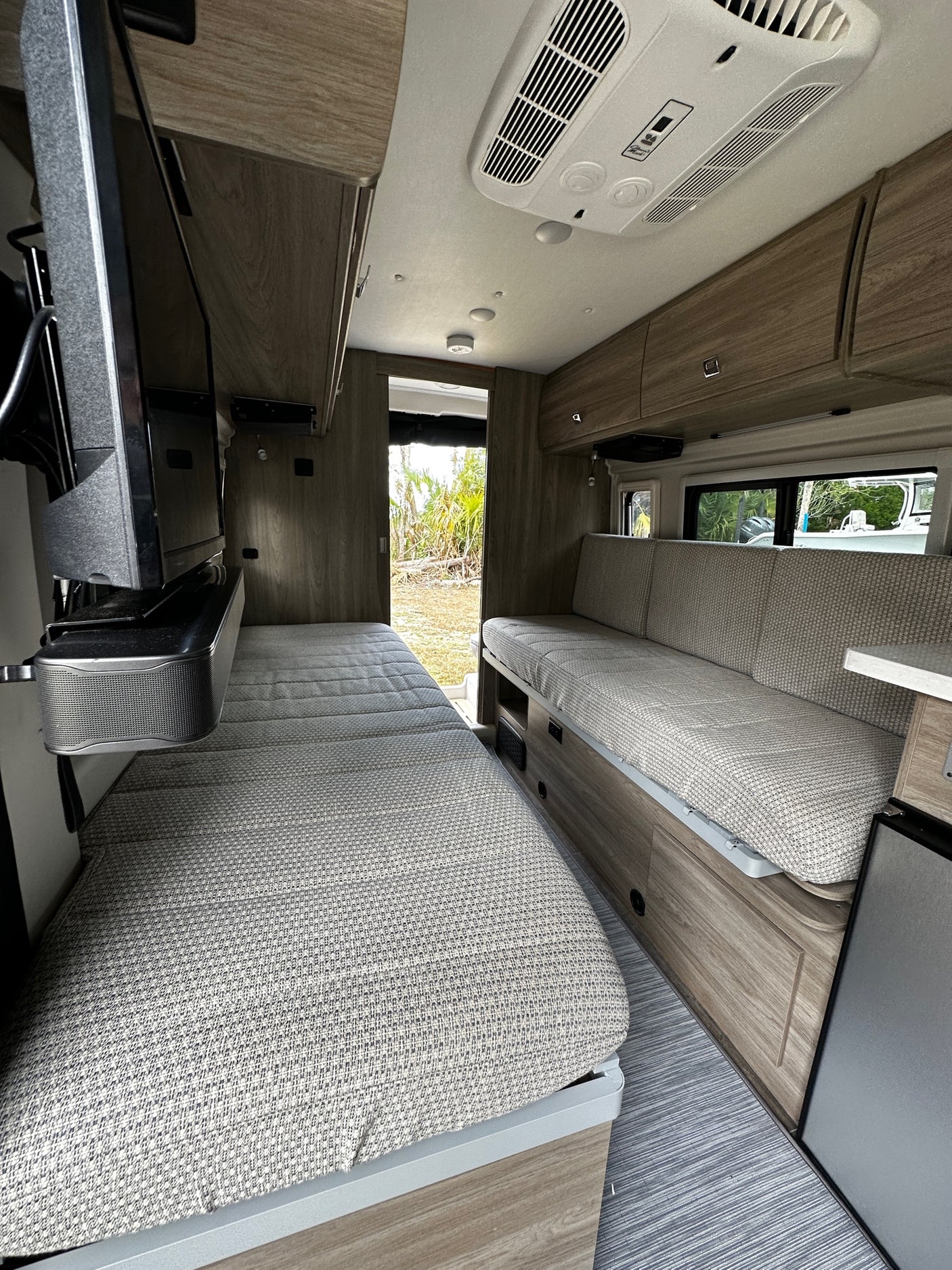 Modern RV rental interior: patterned bed/sofa, wood cabinets, large windows, & ceiling AC unit. Cozy camper space.