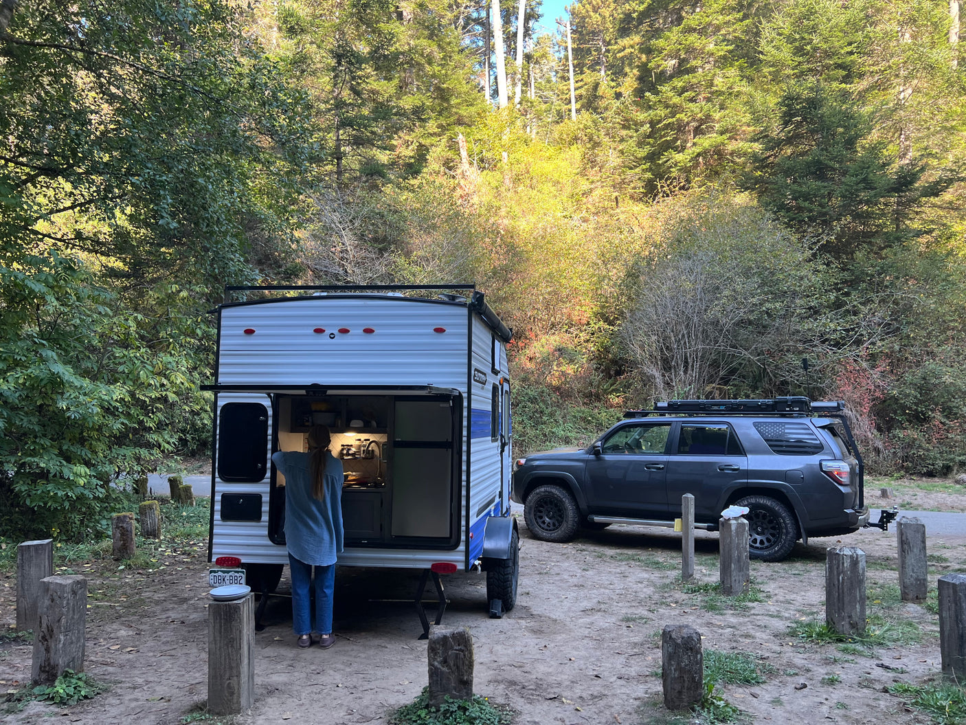 RV rental user prepares food in camper at a wooded campsite, SUV nearby.