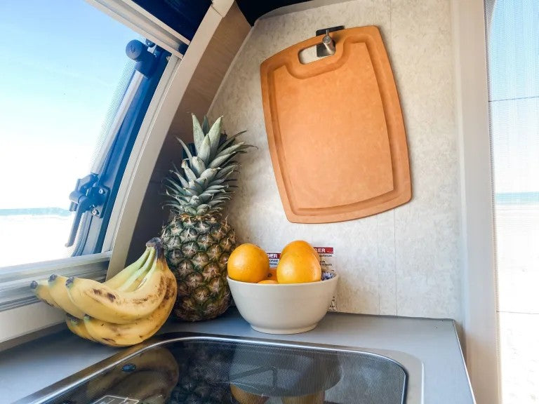 RV rental kitchen with fresh fruit (bananas, pineapple, oranges in a bowl) & cutting board, bright window view of beach.