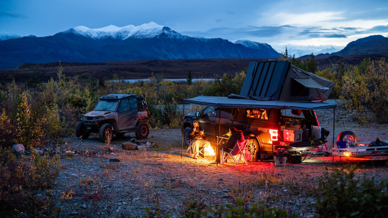 People enjoy a campfire at dusk. An overland truck & UTV offer an RV rental alternative for mountain adventures.