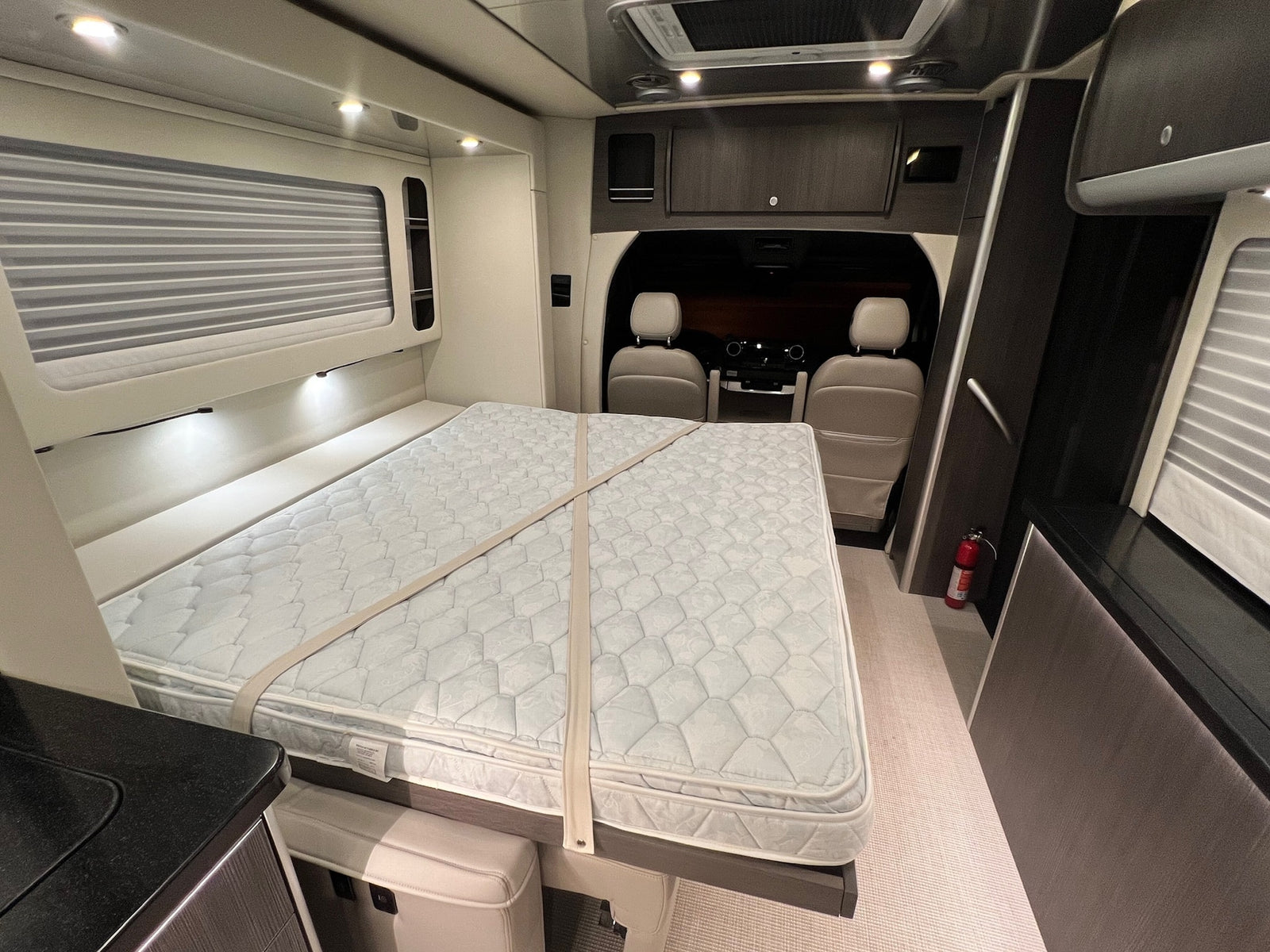 This RV rental interior features a comfy bed with security straps, a fire extinguisher, overhead storage, and front seats for travel.