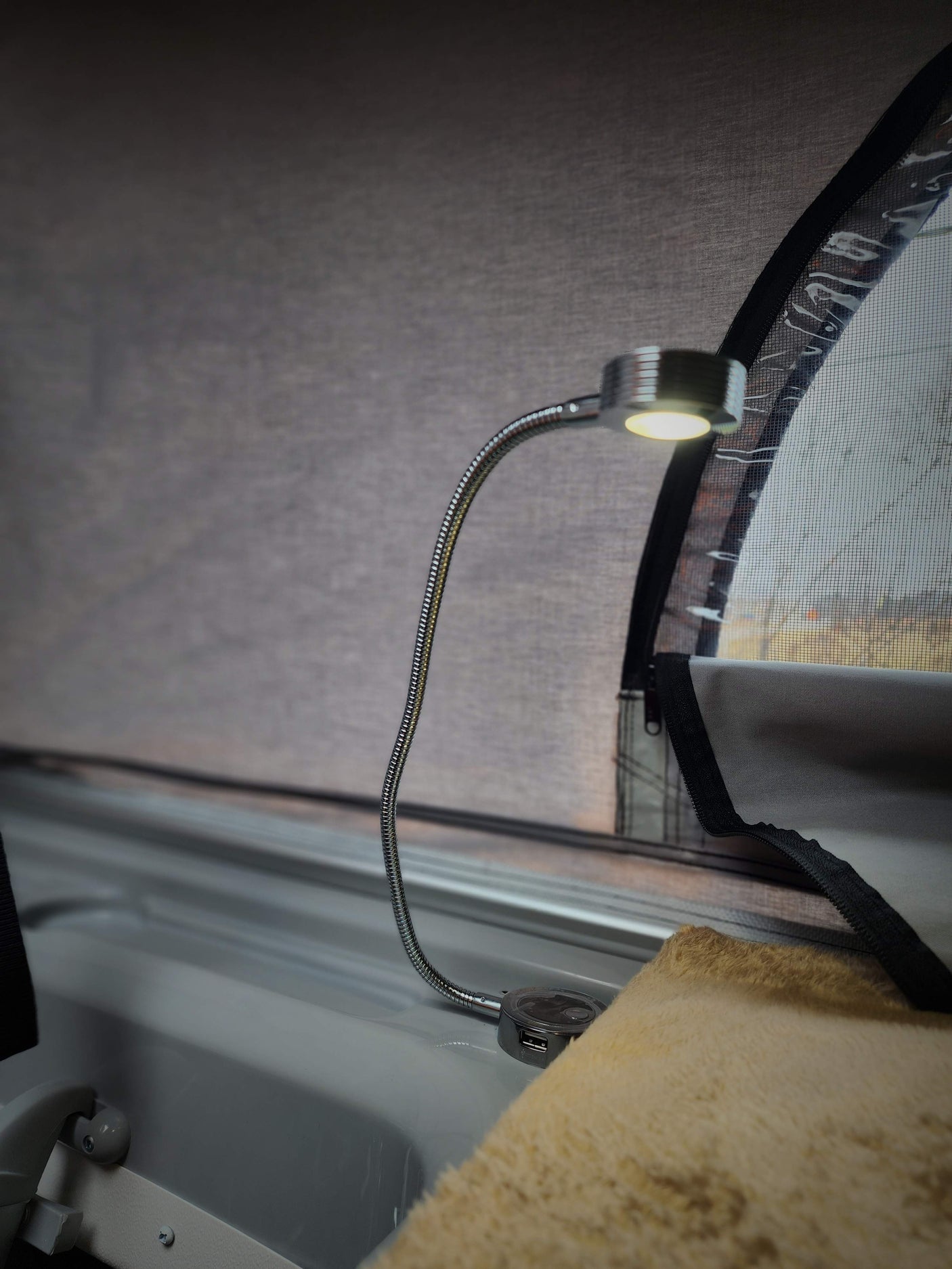 An RV rental's flexible LED light with USB charging on a white panel, beside a furry blanket and window screen.