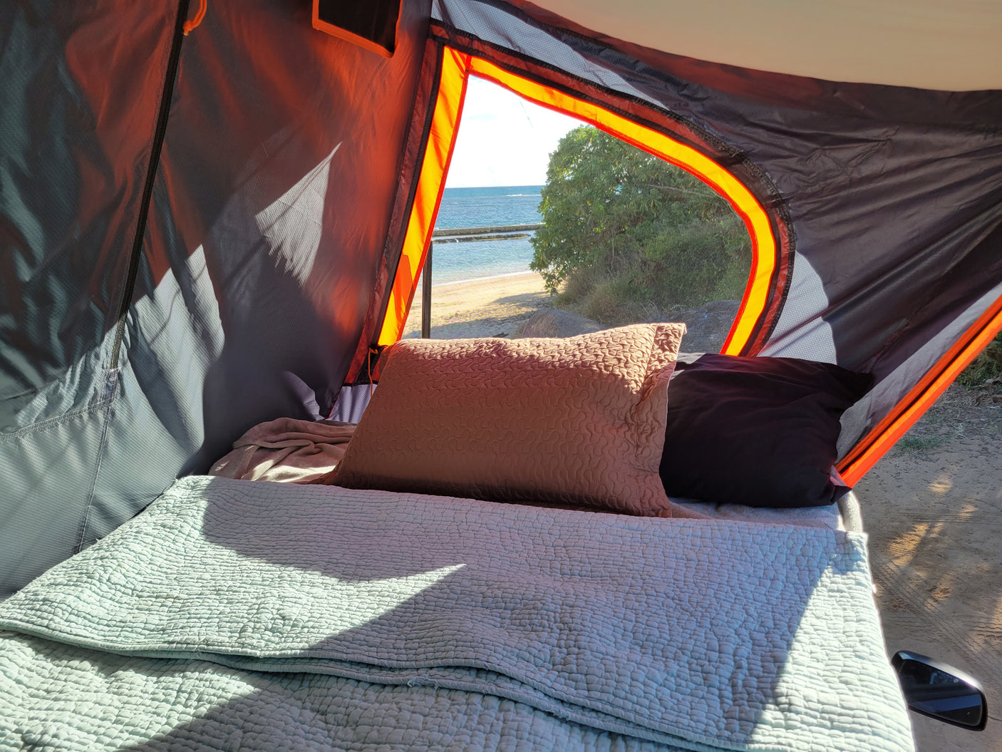 Inside a cozy RV rental, a bed with soft green bedding faces a scenic window offering panoramic views of a sunny beach and ocean.