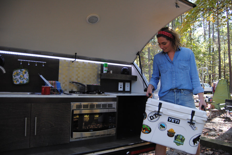 This RV rental features an outdoor kitchen. A woman loads a cooler adorned with National Park stickers, ready for campsite cooking.