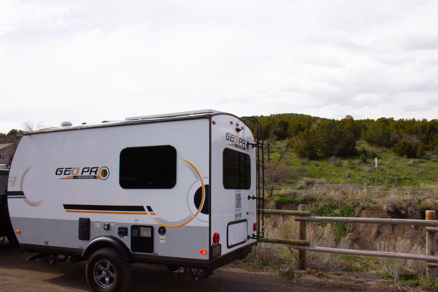 A white and grey GEO PRO travel trailer, available for rv rental, parked outdoors near a fence and trees.