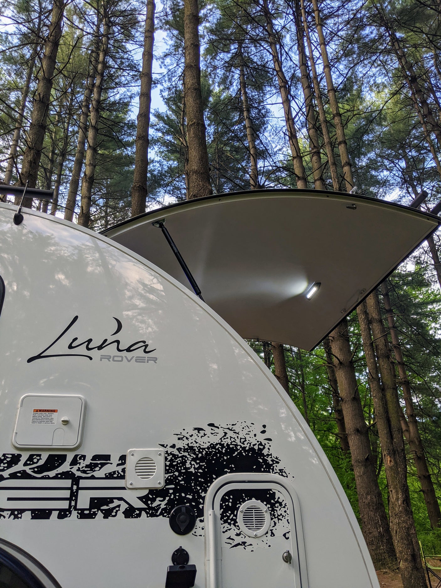 A white Luna Rover teardrop RV rental, with its rear hatch open and lights on, parked in a forest of tall trees for camping.