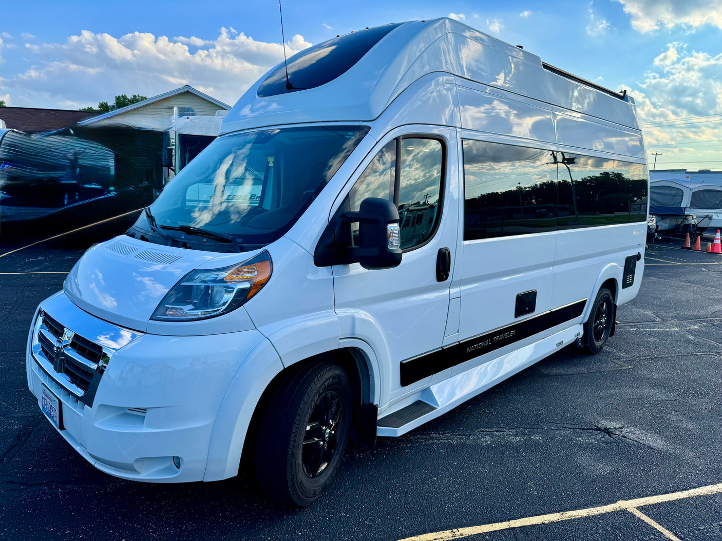 A modern white National Traveler RV rental, with a raised roof and black wheels, is parked on an asphalt lot under a cloudy sky.