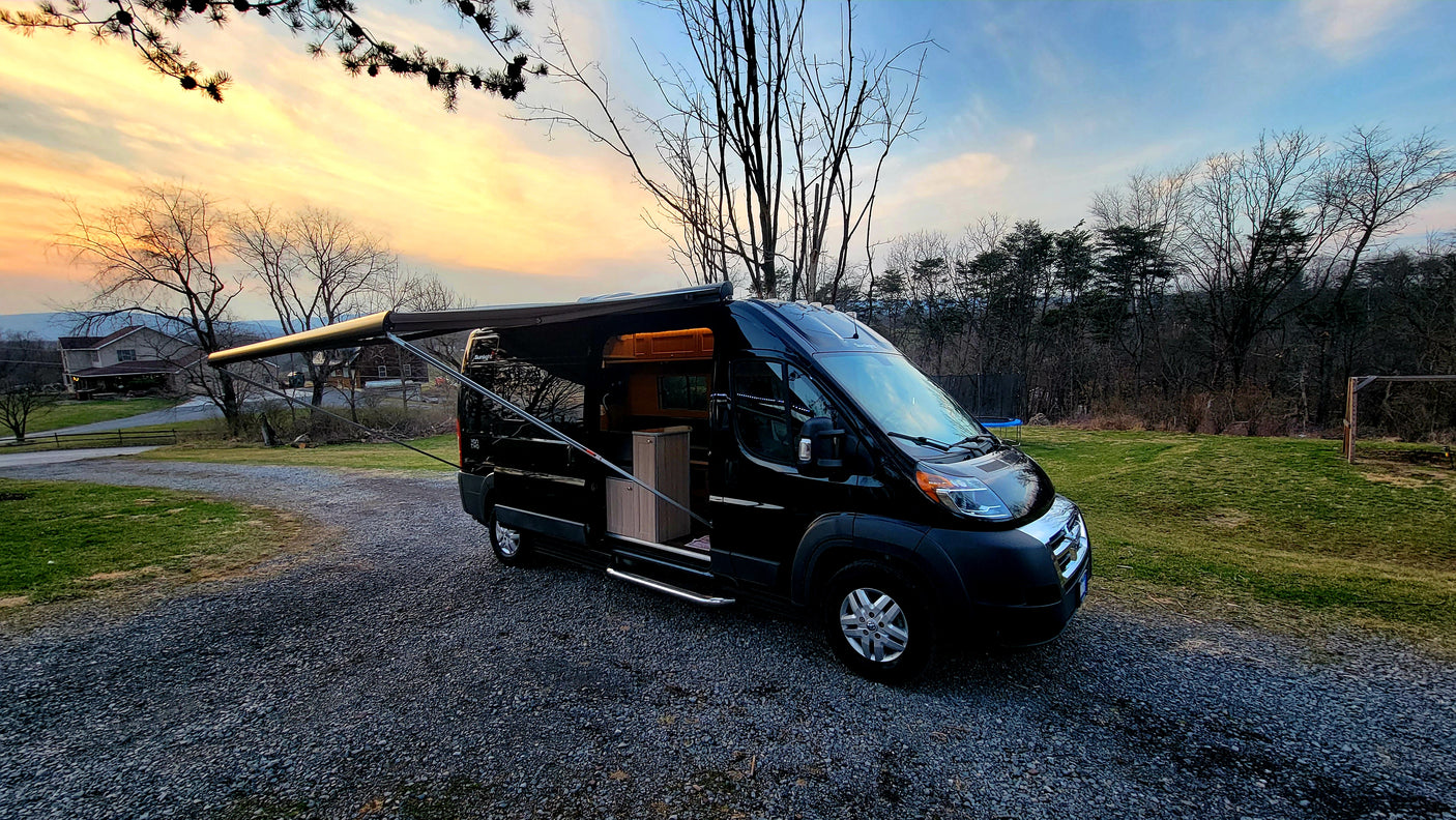 Black camper van for rv rental, parked on gravel with open side door & extended awning, set against a vibrant sunset.