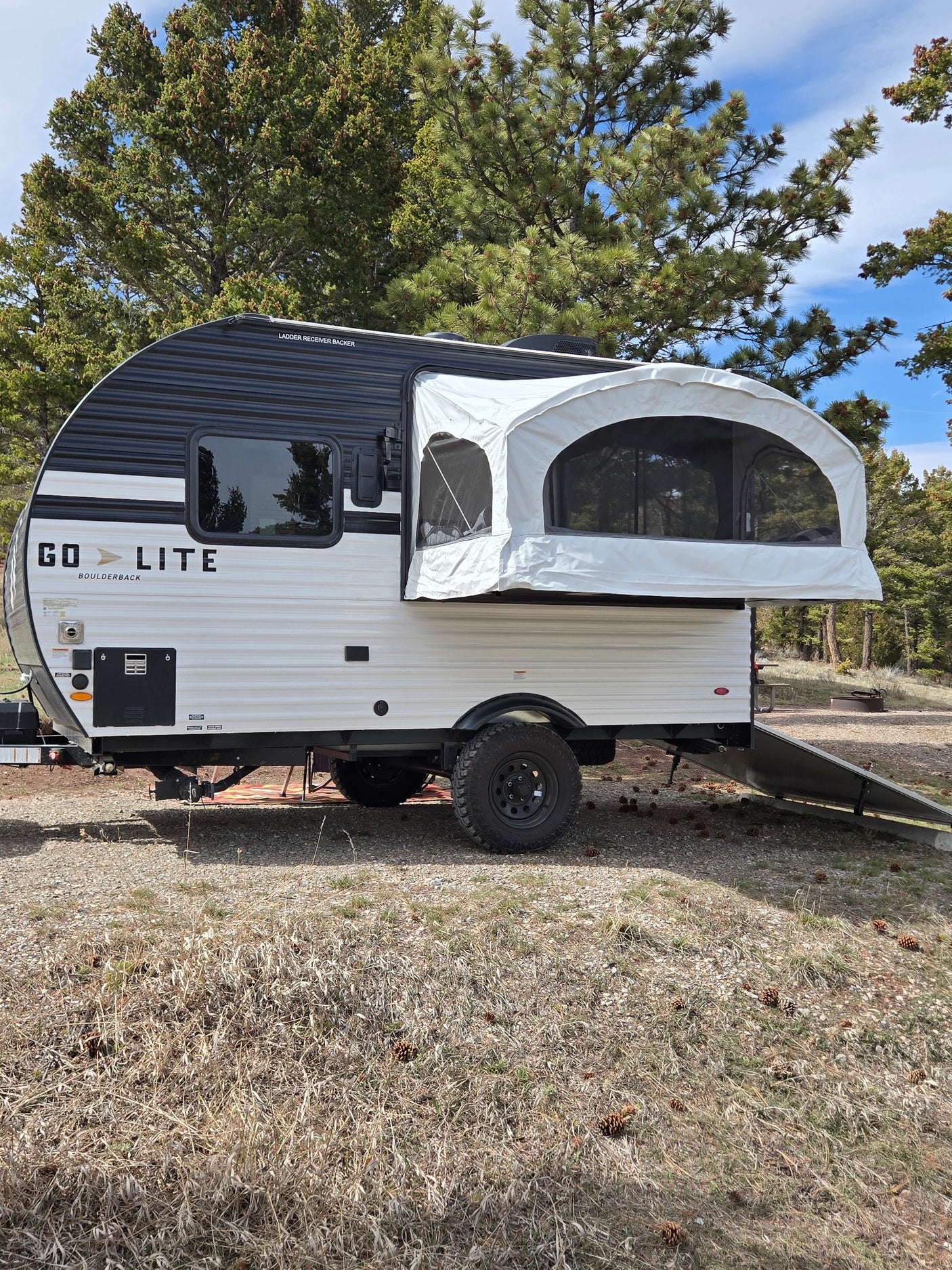 This Go Lite travel trailer rv rental features a pop-out sleeping area, parked on a rustic campsite amidst pine trees.