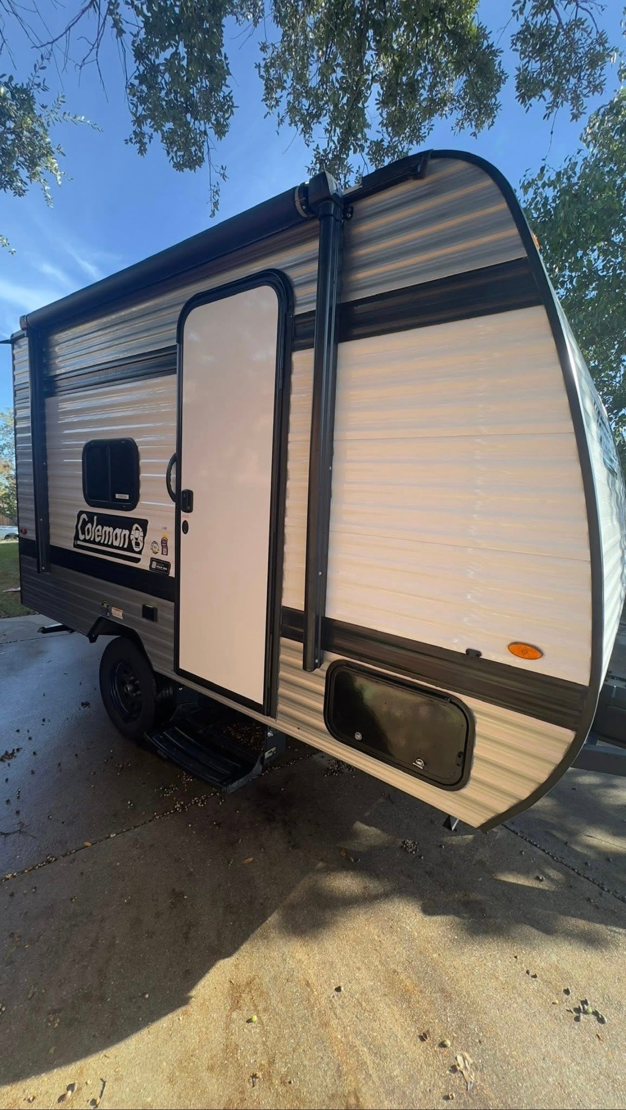 A light-colored Coleman travel trailer RV, ideal for an rv rental, parked on concrete with trees overhead.
