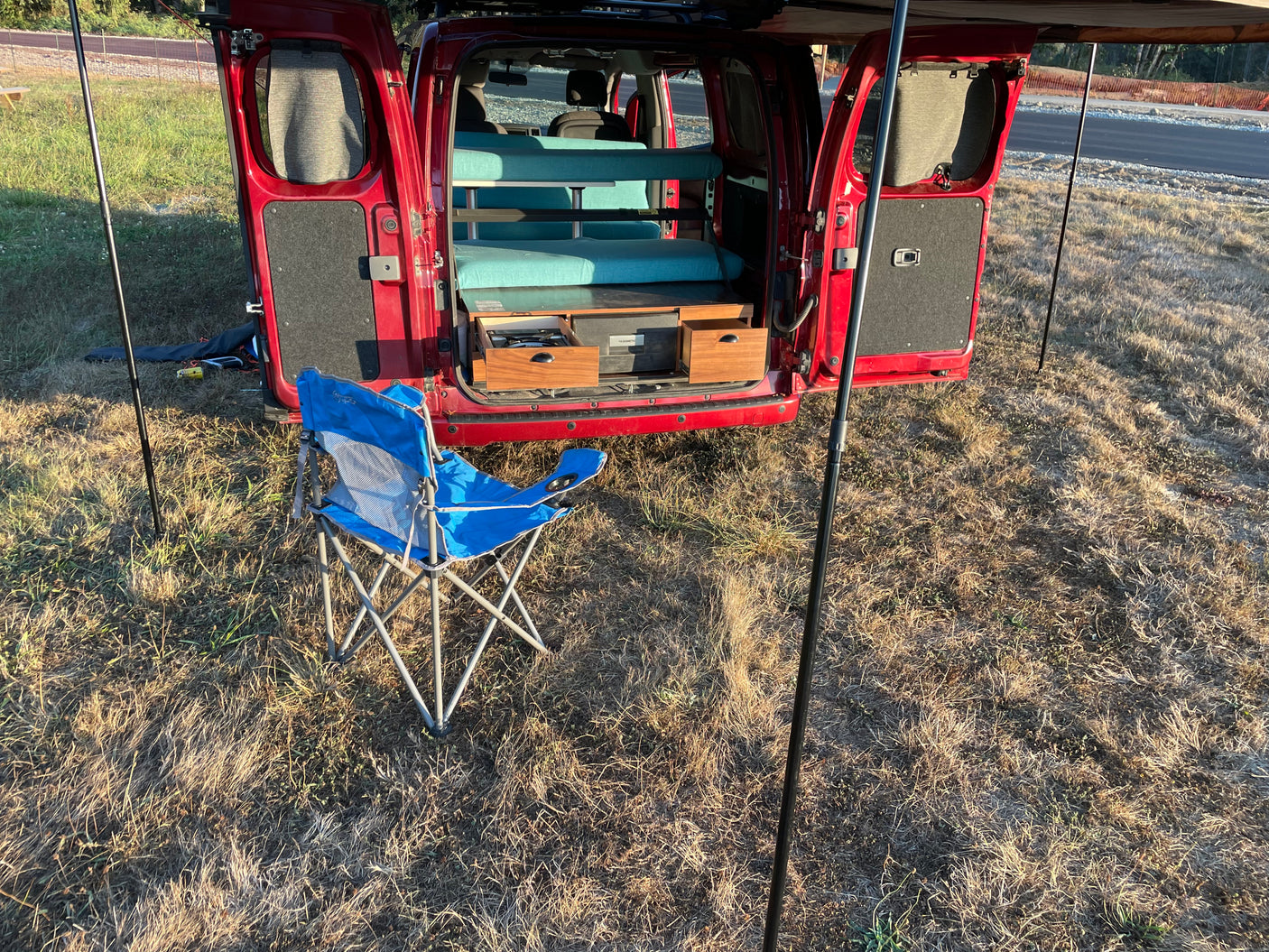 Red camper van with open rear doors reveals a cozy interior bed, pull-out kitchen & a camping chair. Great RV rental setup!