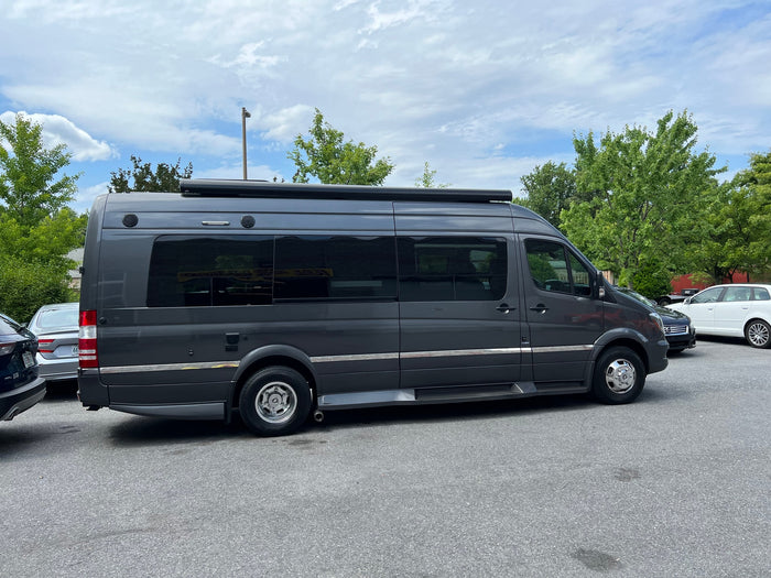 parked on a paved driveway with lush green trees behind it.|A dark grey Mercedes Sprinter camper van