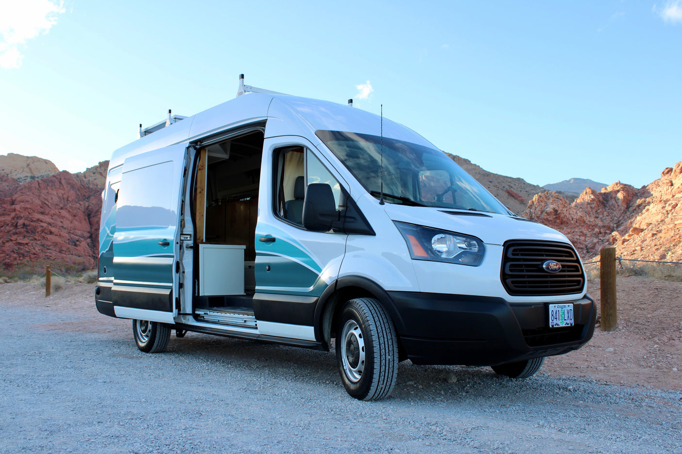 White Ford Transit rv rental camper van on gravel, side door open, amidst red rock mountains under a blue sky.