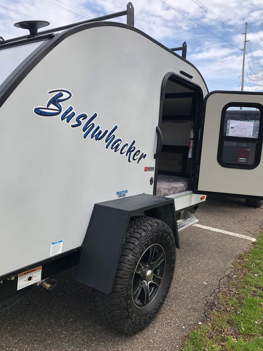 A Bushwhacker teardrop travel trailer for RV rental. Its side door is open, revealing a compact, bed-filled interior.