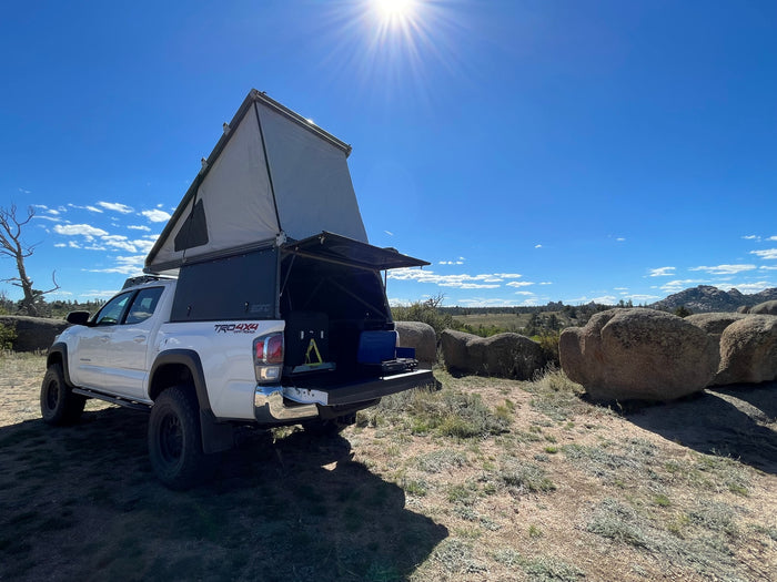 Explore nature with this RV rental! A white Toyota Tacoma truck with a raised rooftop tent & camp kitchen in rocky terrain.
