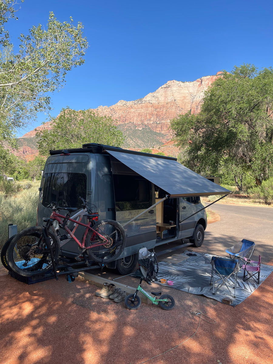 Scenic RV rental campsite: A grey camper van with extended awning, bikes, chairs, and mat, set against stunning red rock mountains.