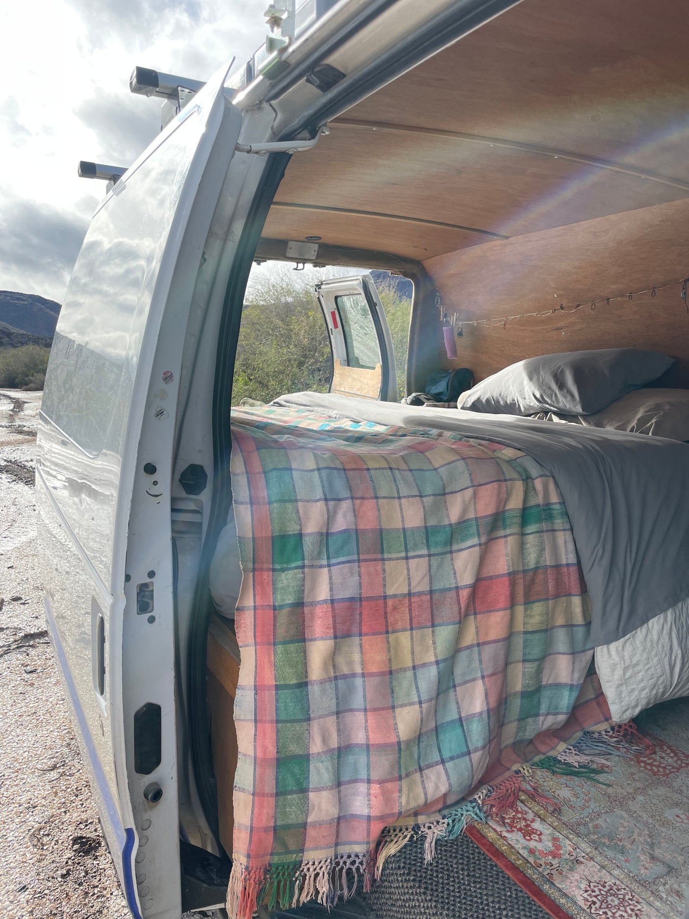 An RV rental camper van's interior: a bed with a colorful plaid blanket, wood ceiling, and open door view of mountains.