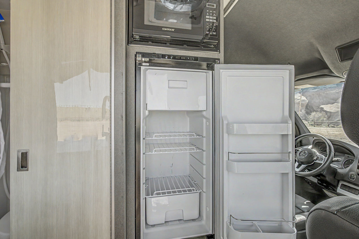 RV rental interior: An open refrigerator and microwave in a camper van's compact kitchen, with a glimpse of the outdoor landscape.