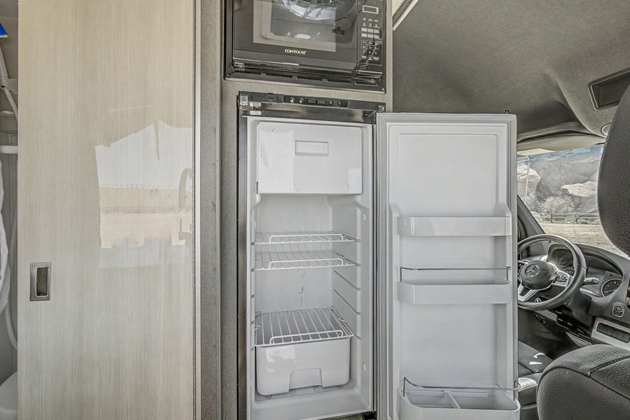 RV rental interior: An open refrigerator and microwave in a camper van's compact kitchen, with a glimpse of the outdoor landscape.
