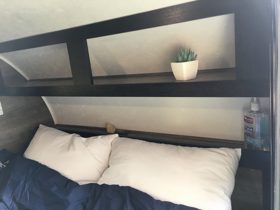 An RV rental's cozy bedroom features a bed with white pillows & blue blanket, plus overhead storage with a plant & Purell.