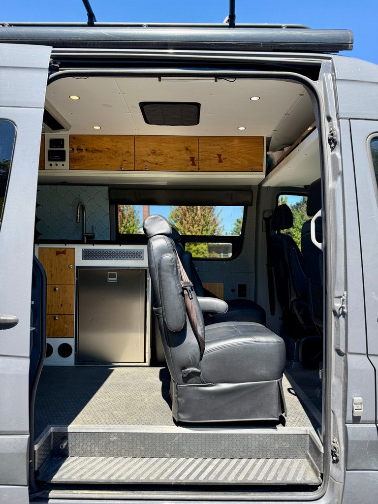 Explore this custom RV rental interior: modern camper van with built-in fridge, wood cabinetry, and swivel seating.