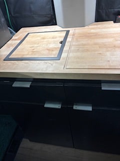 Compact rv rental kitchenette with a wooden countertop featuring a recessed cover, above dark storage drawers and cabinets.