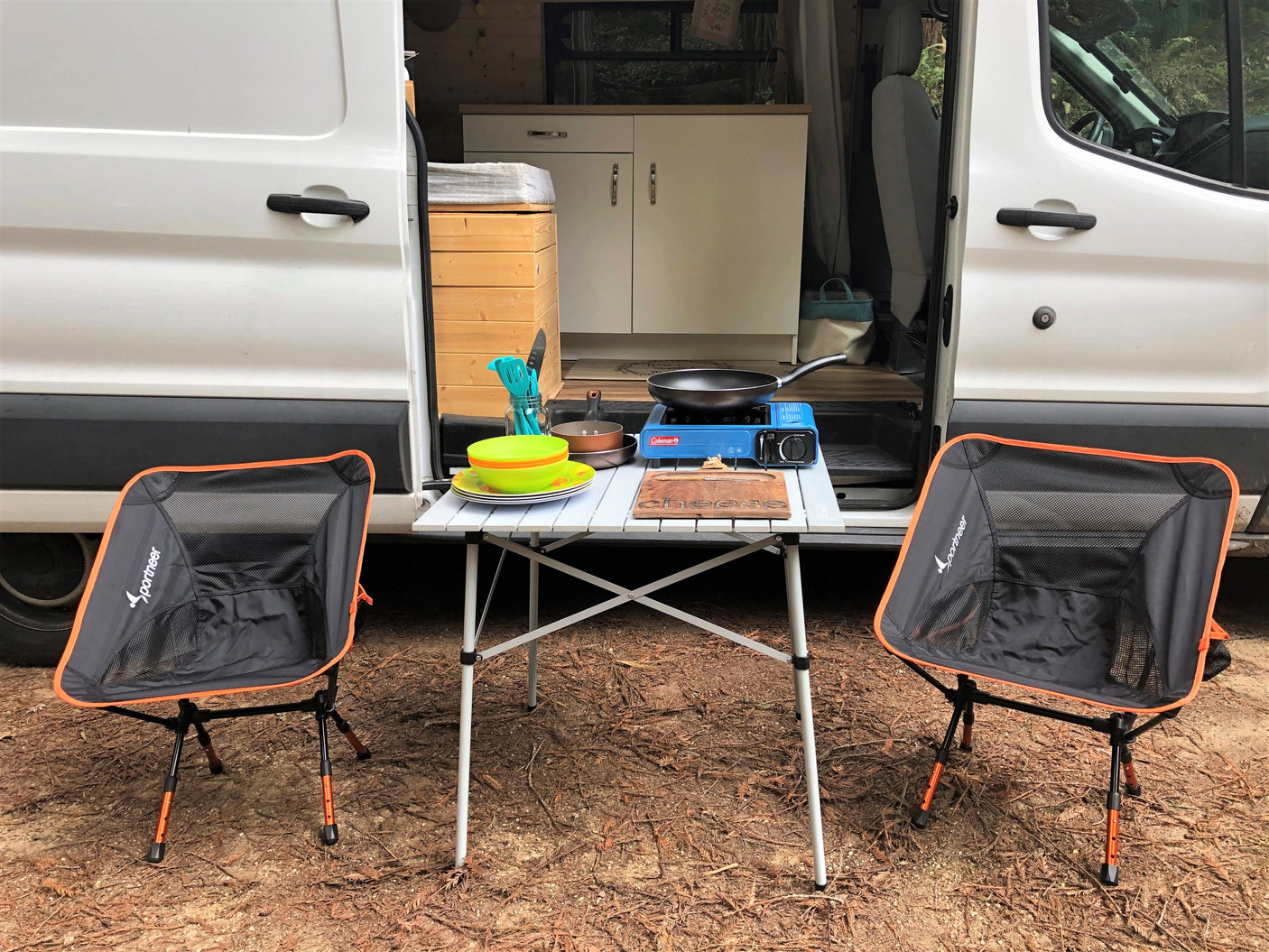 RV rental outdoor kitchen setup: Portable stove, table, and chairs ready for cooking outside a white camper van.