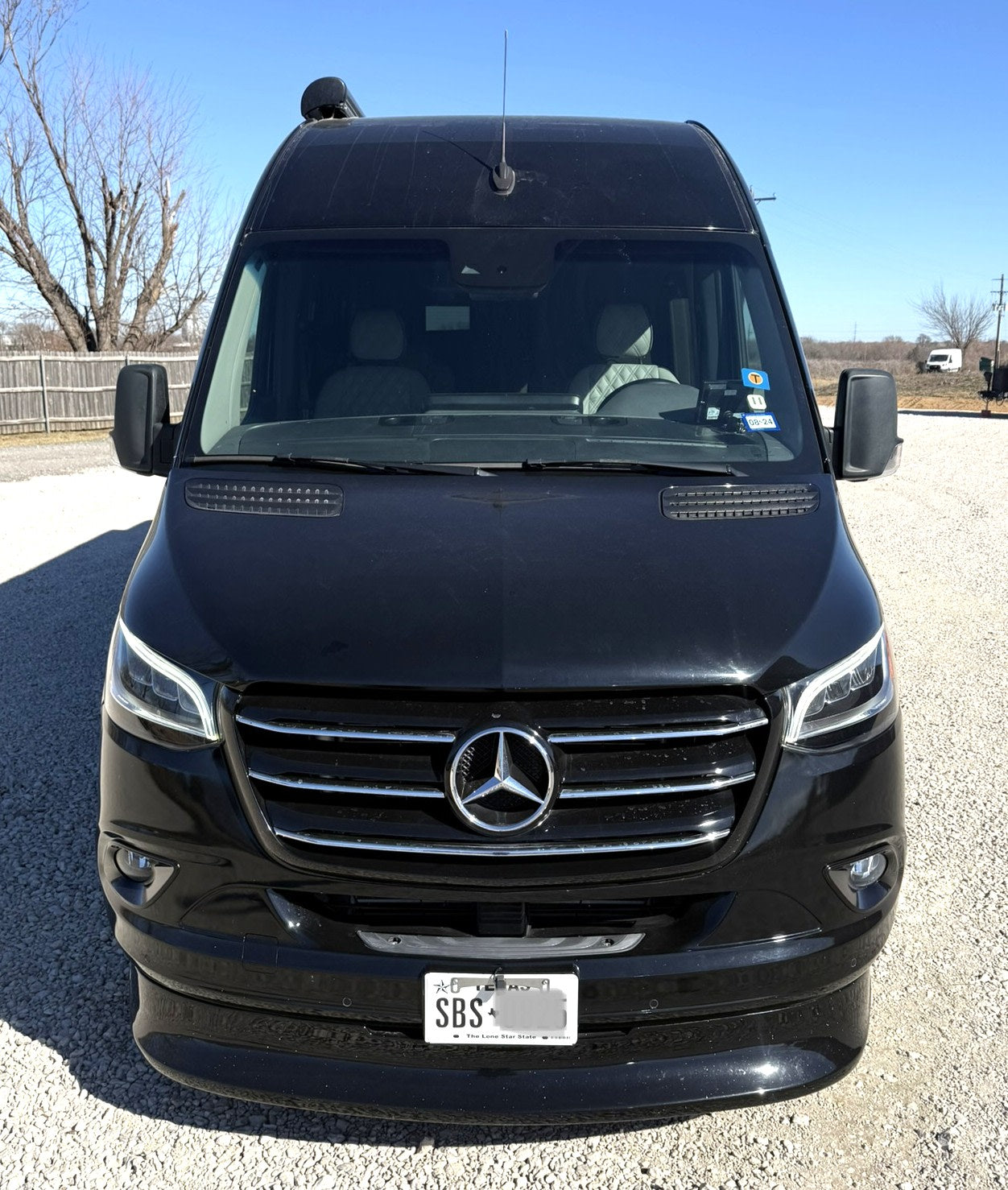 Front view of a sleek black Mercedes-Benz Sprinter van, possibly an RV rental, parked on a gravel lot under clear skies.