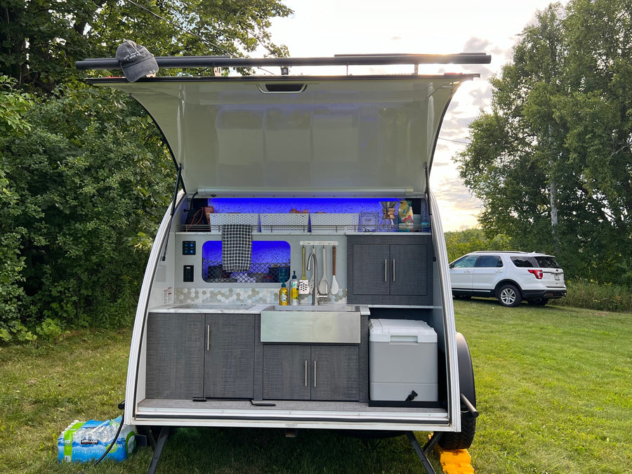parked on grass. Perfect for outdoor camping with barns in background.|Close-up of a Luna Rover teardrop rv rental with black splatter graphics