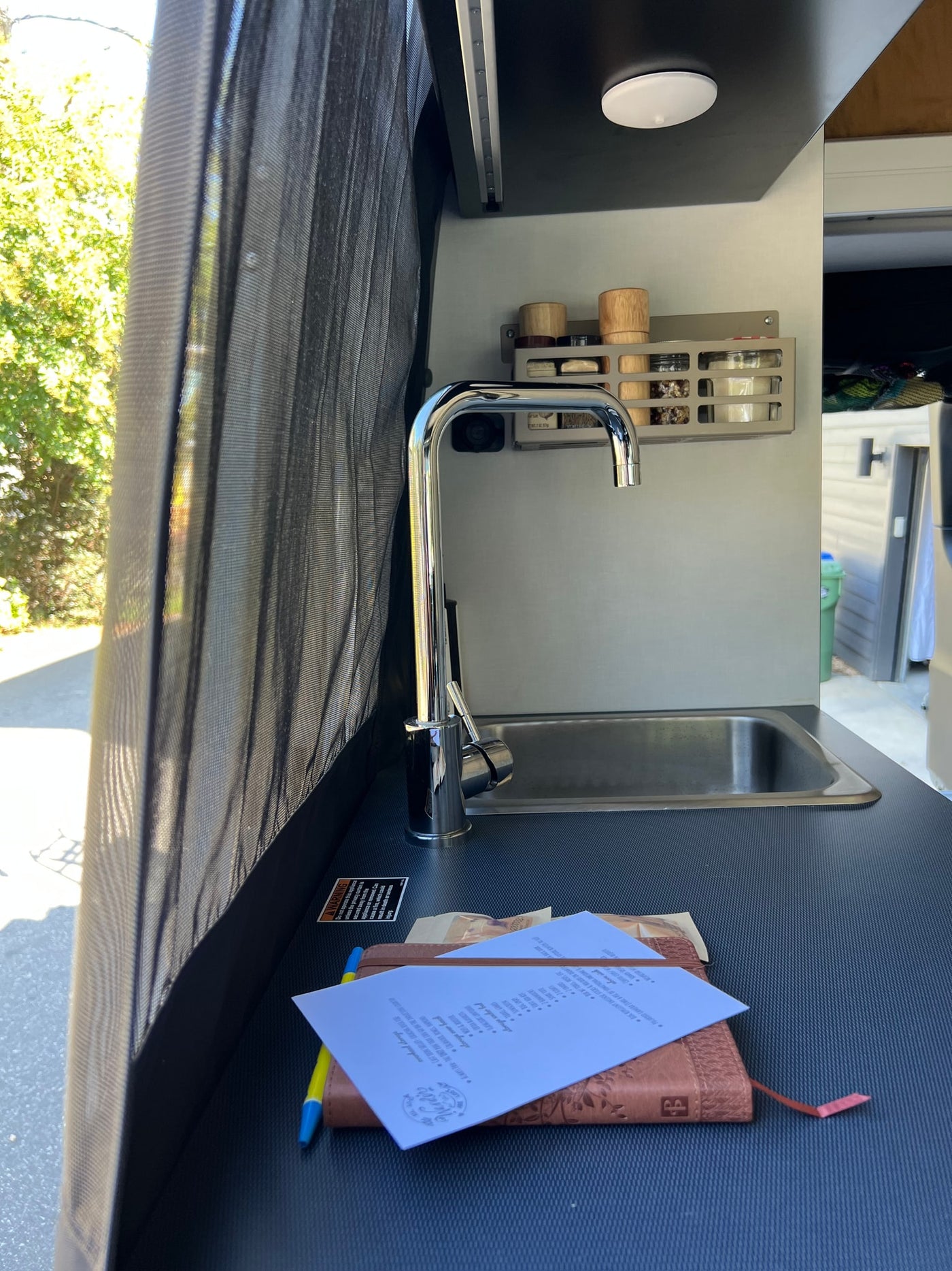 RV rental kitchenette featuring a sink, modern faucet, and spice rack. A warning label and journal are on the counter. — photo 18