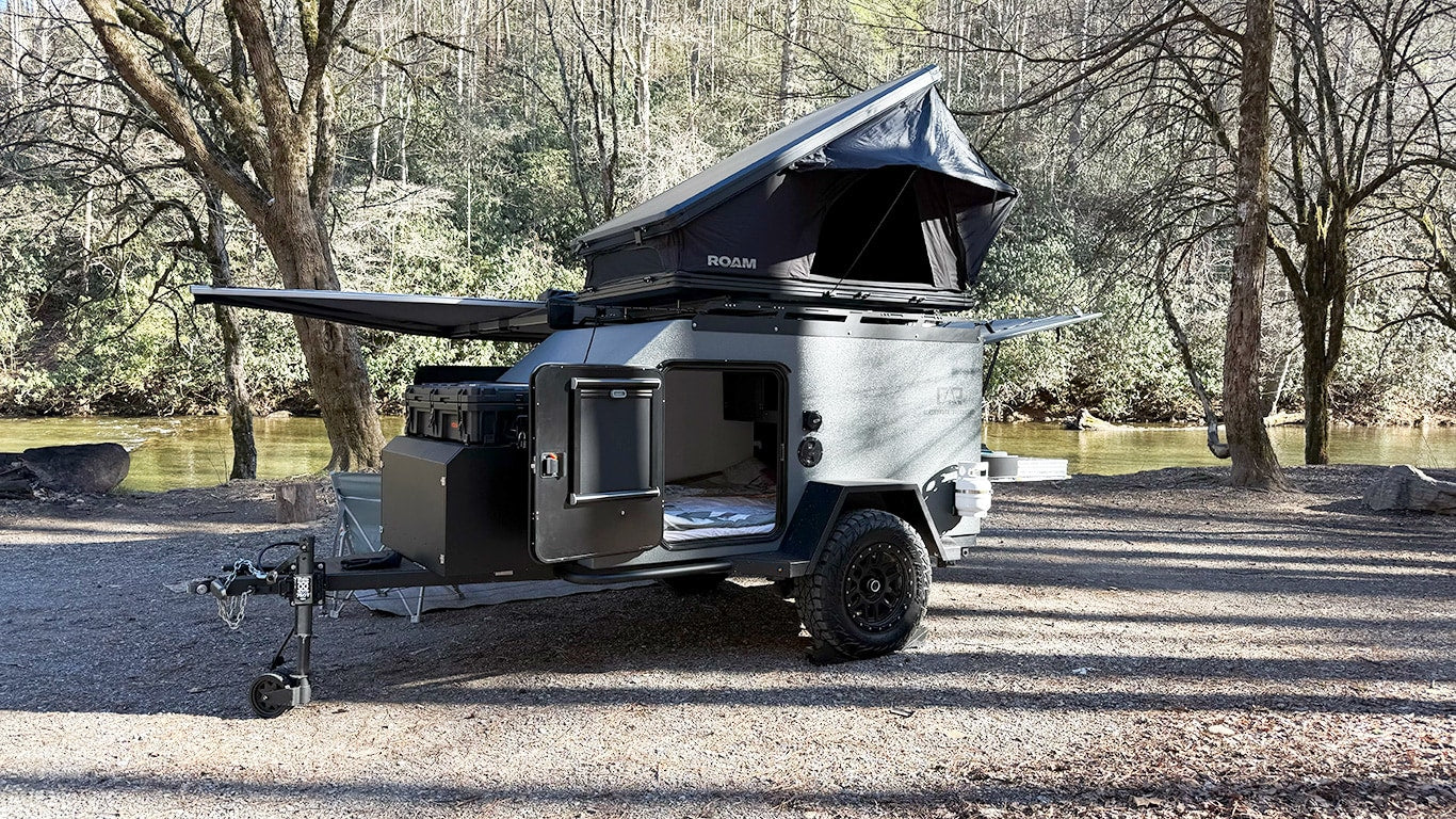 An off-road camper trailer RV rental with a rooftop tent and awning is parked by a scenic river in the woods.|Rugged off-road camper trailer with a rooftop tent and awning