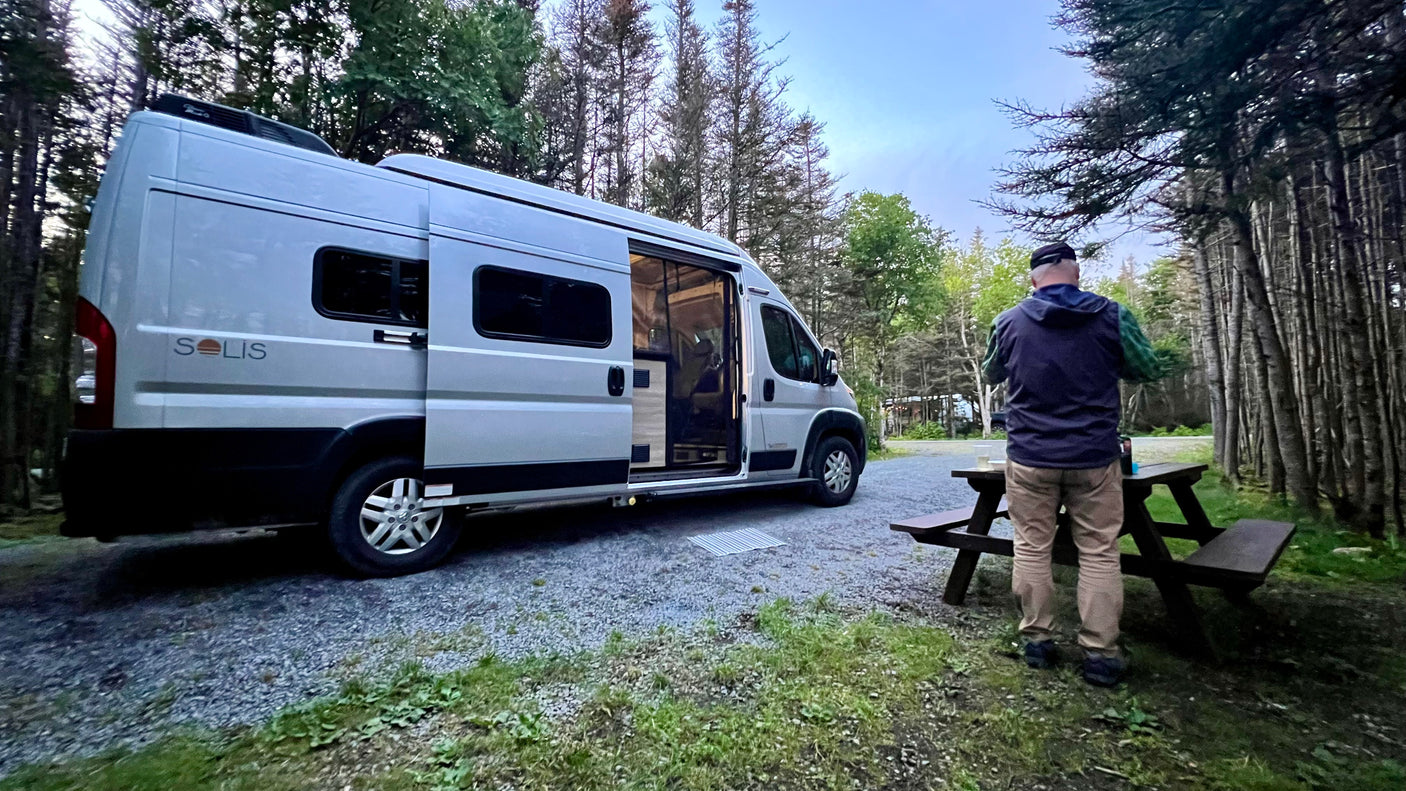 An RV rental, a silver Winnebago Solis, parked in a tranquil forest campsite at dusk. A person stands near a picnic table.