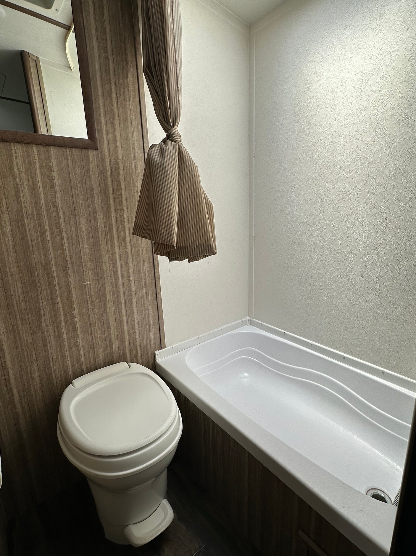 Clean RV rental bathroom featuring a compact toilet, bathtub, and tied-back shower curtain. Wood-paneled walls and a light-colored shower surround are visible.