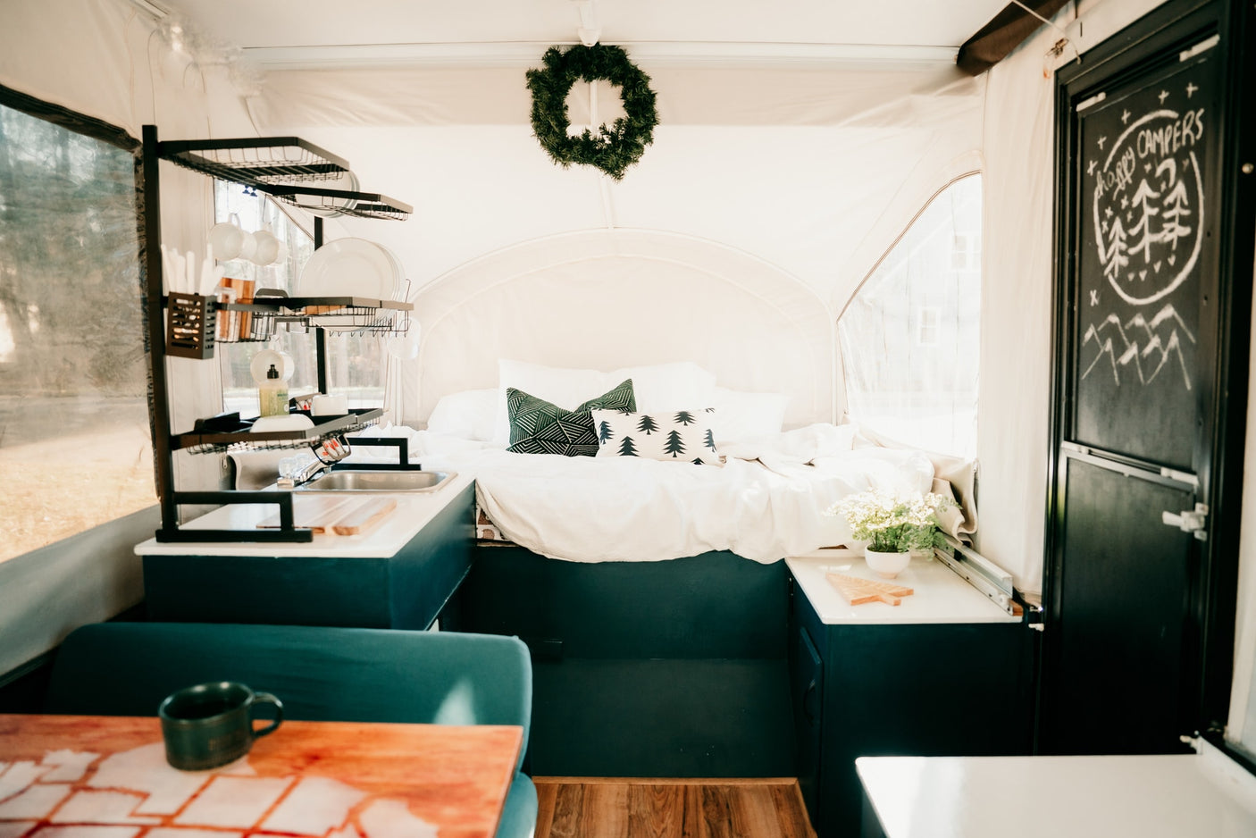 Cozy RV rental interior featuring a comfy bed, compact kitchen with sink, and a "Happy Campers" chalkboard door.