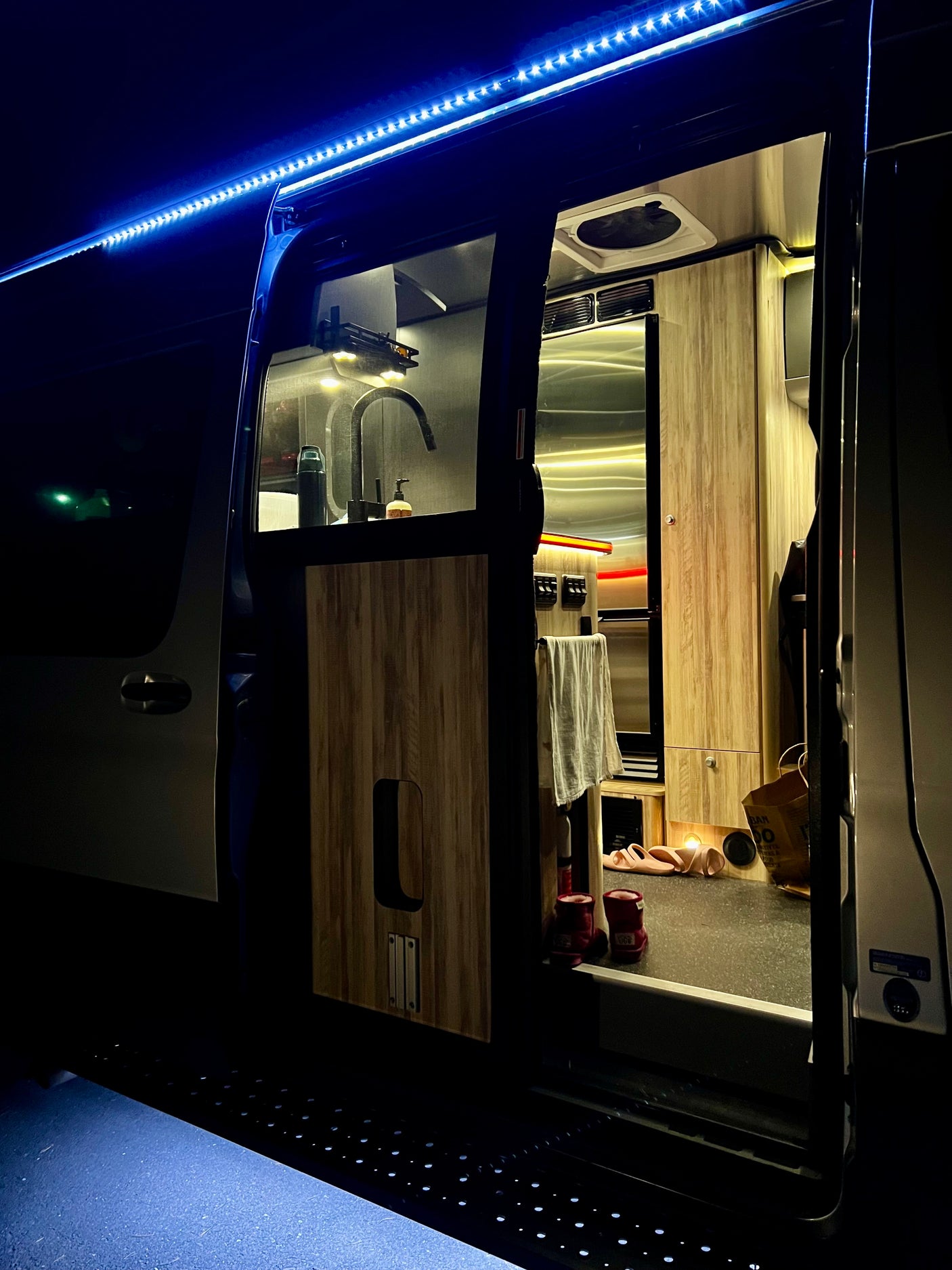 Cozy rv rental interior at night, featuring bright blue exterior LEDs, a lit kitchen, and an open camper van side door.