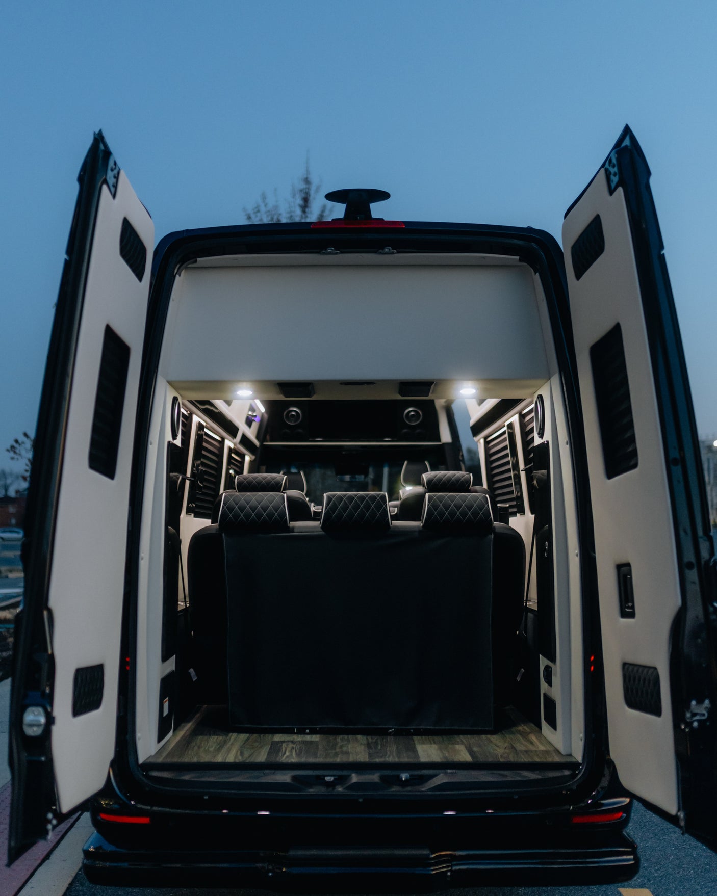 Open rear doors reveal a premium RV rental's spacious interior: comfortable quilted seating, wood flooring, and lighting.