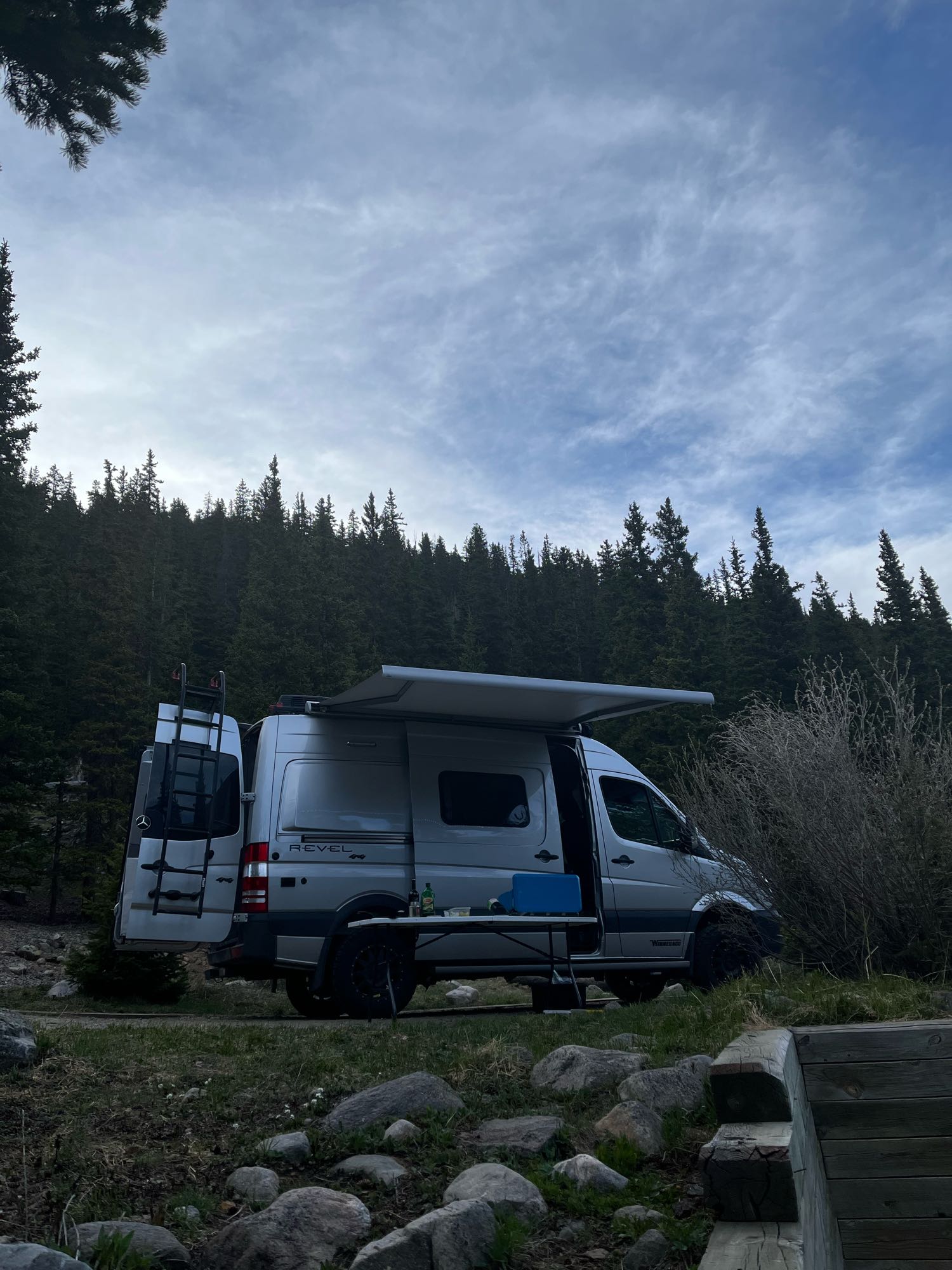 with green forested slopes behind.|Silver Winnebago Revel camper van